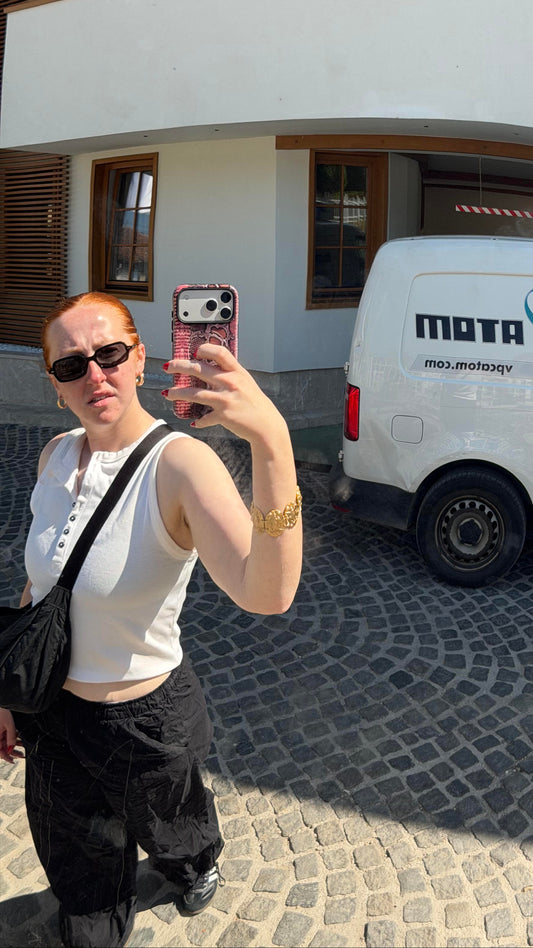 A woman taking a mirror selfie in front of a white building and a white MOTA delivery van parked on a cobblestone driveway. The image is framed by a red curved border, likely a mirror edge.